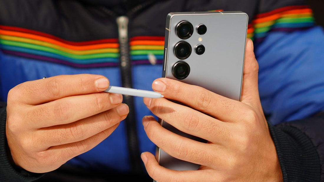 A close-up of a person holding a silver Samsung Galaxy Ultra smartphone in one hand and a white stylus in the other, wearing a blue and black puffer jacket with a rainbow stripe across the chest.