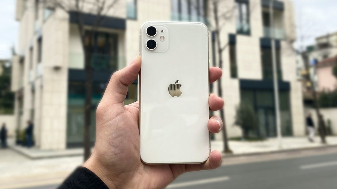 A hand holding an Apple iPhone with a white backplate and gold logo, photographed outdoors with a blurred city street and modern building in the background.