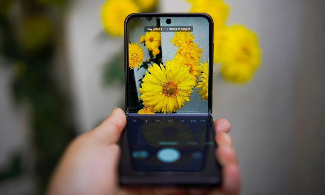A hand holding a black foldable smartphone showing a camera app screen with a yellow flower photo, against a blurred background of yellow flowers and green foliage.