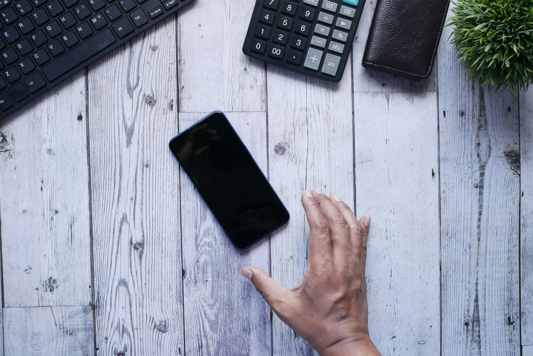 Hand reaching towards a smartphone on a wooden desk with a keyboard, calculator, and wallet in the background. lost android phone