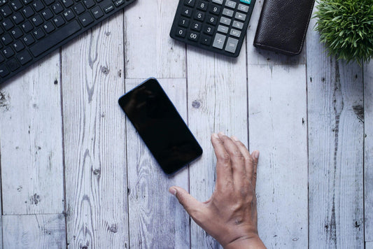 Hand reaching towards a smartphone on a wooden desk with a keyboard, calculator, and wallet in the background. lost android phone