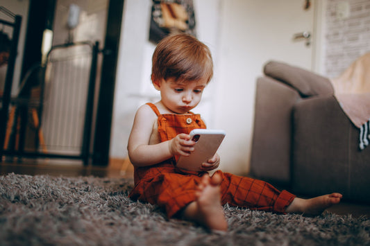 Child, toddler, in an orange outfit playing with a tablet on a carpeted floor.