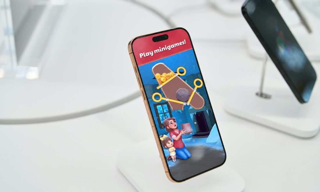 A smartphone with a gold-toned edge stands on a white display stand, showing a colourful app/game screen Gardenscapes with the text 'Play minigames!'. A second phone on a stand is blurred in the background on a white surface.